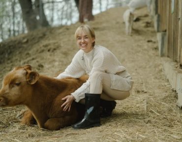 Smiling woman wearing boots joyfully interacting with a calf outdoors on a rural farm path.