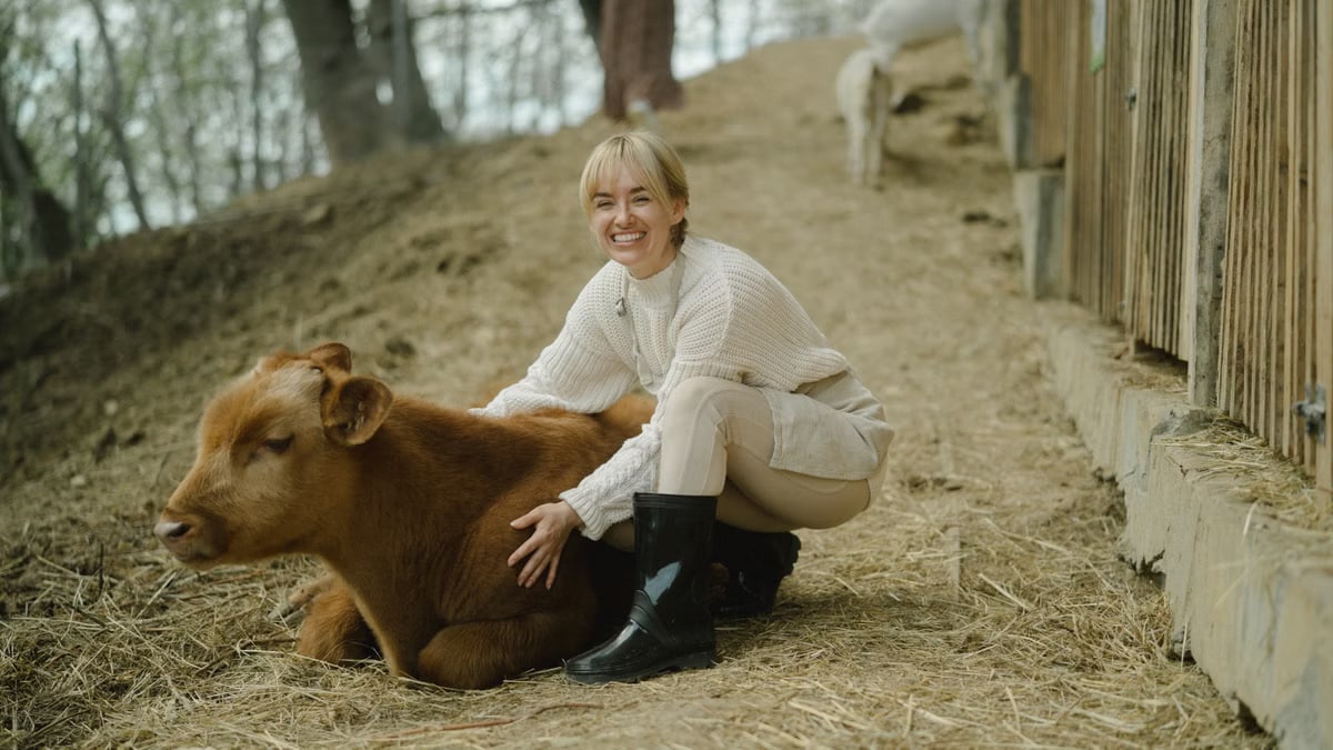 Smiling woman wearing boots joyfully interacting with a calf outdoors on a rural farm path.