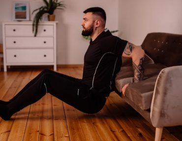 A bearded man in black fitness attire exercising at home, showcasing arm tattoos and a healthy lifestyle.